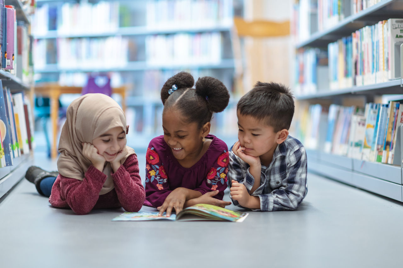 Three children of diverse cultural background lying on the floor of a library reading a book together